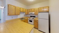 This single-floor unit offers a functional and straightforward layout connecting the main living areas seamlessly. The open-plan living room and kitchen form the heart of the space, with neutral-toned walls and tan wall-to-wall carpeting throughout the living and bedroom areas. Exposed ceiling beams add a subtle architectural detail to the living room, which flows directly into the kitchen via a peninsula counter.

The kitchen is well-equipped, featuring warm wood-toned cabinetry extending to the upper walls for ample storage, light laminate countertops, a double-coil electric range, a refrigerator, an under-cabinet range hood, and a single-basin sink. Resilient flooring in the kitchen provides a practical contrast to the carpeted living areas.

The bedrooms are generously proportioned, each benefiting from large multi-pane windows that draw in natural light and offer outdoor views. Both rooms include baseboard heating and bifold-door closets with built-in rods for organized storage.

The bathroom is clean and efficiently designed, featuring a combined tub and shower with a handheld showerhead, a toilet, and a vanity with a wood-toned cabinet base, countertop sink, and a lighted mirror above.