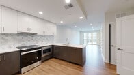 This thoughtfully designed single-floor residence showcases a contemporary open-concept layout that seamlessly connects the kitchen and living areas. The kitchen is a standout feature, offering two distinct design configurations: one with a two-tone aesthetic of white upper cabinetry and dark espresso lower cabinets paired with a peninsula sink, and another with striking blue lower cabinetry and a full island. Both configurations feature stainless steel appliances, a marble-style tile backsplash, and clean white countertops that enhance the modern aesthetic. Recessed lighting throughout maintains a bright, airy atmosphere. Warm-toned hardwood flooring runs continuously across the main living areas, adding a sense of cohesion and natural warmth. The living area is spacious and open, with large sliding glass doors leading to a private balcony with courtyard views, flooding the space with natural light. A dedicated bedroom features a generously sized multi-panel window overlooking a landscaped communal courtyard. The bathroom is finished with floor-to-ceiling marble-patterned porcelain tile, a soaking tub with an overhead shower, a floating vanity, and chrome fixtures, creating a clean and polished spa-like feel throughout.