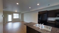 This three-floor space showcases a thoughtfully designed open-concept main living area that seamlessly integrates the kitchen and living room. The kitchen is a standout feature, anchored by a dark-toned island with a double stainless steel sink and rich granite-style countertops. Dark cabinetry complements a suite of black appliances, including a French door refrigerator and range, creating a bold, cohesive aesthetic. Recessed lighting is distributed across the ceiling, providing consistent, bright illumination throughout the open area. The living space is generously proportioned, offering ample room for a variety of furniture arrangements. Warm hardwood-style flooring flows continuously throughout, adding a welcoming feel underfoot. Neutral-toned walls contribute to a clean, versatile backdrop suited to many décor styles. A window along one wall allows natural light to filter in, brightening the space. Visible HVAC vents indicate a central air system for year-round comfort. The overall design balances modern finishes with functional layout across all three floors of the property.