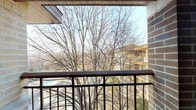 This single-floor residential unit offers a practical and comfortable layout designed for everyday living. The open living area is finished with wall-to-wall carpet and neutral white walls, creating a clean and versatile space. A sliding glass door leads directly to a private balcony, where a metal railing frames an elevated view of mature trees and the surrounding neighborhood, providing a pleasant natural backdrop.

The kitchen is efficiently arranged in an L-shaped configuration, featuring warm honey-toned wood cabinetry, white appliances including a freestanding range and full-size refrigerator, a double-basin sink, and light-toned tile flooring. Overhead fluorescent lighting ensures a well-lit workspace throughout.

The bedroom is generously proportioned with soft carpeting and includes a built-in closet with sliding panel doors and a full-width hanging rod for ample storage. A hallway connects the bedroom to the main living spaces, and the front entry features a solid wood door with carpet continuing into the foyer area.

Throughout the unit, warm wood-toned trim and baseboards provide consistent detailing, while multiple vents indicate a central HVAC system. The layout is straightforward and functional, offering a comfortable living environment with the added benefit of outdoor access and natural light through the balcony door.