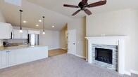 This single-floor home showcases a thoughtfully designed open-concept layout that connects the living area and kitchen in a cohesive, functional flow. The living room is anchored by a classic white-mantled fireplace with a stacked stone surround, adding a refined focal point to the space. Plush carpeting underfoot lends warmth and comfort to the area, while a ceiling fan with a dark wood finish overhead contributes to both the aesthetic and practical functionality of the room. Vaulted ceilings rise above, creating an airy, expansive atmosphere enhanced by recessed lighting throughout. The kitchen area features a granite-topped peninsula with pendant lighting hanging above, offering a casual dining or workspace option. Stainless steel appliances are visible from the living space, reinforcing the kitchen's modern, well-appointed character. Neutral, warm-toned walls tie the spaces together seamlessly, while durable hardwood-style flooring in the kitchen and transition areas complements the soft carpeting in the living zone. A crisp white interior door leads to additional areas of the home, maintaining a clean and cohesive design language throughout. The overall ambiance is welcoming, bright, and move-in ready.