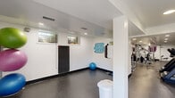 This fitness center is a thoughtfully designed, single-floor workout facility that provides a complete exercise environment within a generously sized open layout. The space is divided into two functional zones: a dedicated stretching and floor exercise area near the entrance, and a main equipment room extending toward the rear.

The entry zone features durable rubber flooring, white walls, and natural light from ground-level windows. A yoga mat, stability ball, and resistance accessories are neatly stored here, alongside a bulletin board displaying exercise guides and workout charts. Motivational wall graphics — including an ab challenge poster — add an energetic atmosphere throughout.

The main equipment area is lined with large wall mirrors that enhance the sense of depth and allow users to monitor their form. Recessed lighting illuminates the space evenly. Cardio equipment includes treadmills, elliptical trainers, and a stationary bike, while the strength training section features cable machines, a functional trainer, and a weight bench. A wall-mounted television provides entertainment during workouts.

The space is finished with dark speckle rubber flooring throughout, white walls, and black base trim, creating a clean, professional aesthetic. A dedicated exit door with signage ensures safe egress. Overall, the facility is well-maintained and equipped to support a wide variety of fitness routines.
