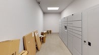 This package room is a purpose-built space designed to efficiently manage incoming deliveries. The room is finished with clean white walls and durable polished aggregate flooring, creating a bright and functional environment. Recessed flush panel lighting in the ceiling provides consistent, even illumination throughout the space.

A prominent bank of multi-unit smart parcel lockers lines one wall, offering secure, individual compartments in various sizes to accommodate packages of differing dimensions. These lockers provide a reliable and organized solution for parcel storage and retrieval.

The room offers generous open floor space, allowing for the temporary placement and sorting of larger deliveries and oversized parcels that may not fit within the standard locker compartments. A secondary door provides additional access, supporting smooth logistical flow for deliveries and pickups.

Security is reinforced by overhead surveillance cameras positioned to monitor the full extent of the room. The overall design is utilitarian yet well-maintained, prioritizing practicality and ease of use. This package room serves as a centralized, secure hub for all incoming shipments, ensuring convenient and organized parcel management.