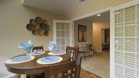 This single-floor residence offers a welcoming and functional layout designed for comfortable everyday living. The dining area serves as a central gathering space, featuring a classic round wooden table with seating for four, complemented by warm neutral-toned walls and hardwood flooring that adds a touch of timeless charm. Decorative wall art, including an eye-catching arrangement of colorful plates, enhances the character of the space and contributes to its homey ambiance. Natural light filters in through plantation-style shuttered windows, lending a bright and airy feel to the interior. Just beyond the dining area, a cozy sitting nook with a petite bistro table and chairs provides an additional intimate corner, ideal for morning coffee or casual conversation. Recessed lighting throughout ensures consistent, comfortable illumination across the space. The layout flows seamlessly from the dining and living areas toward the bedroom, which is visible through a connected hallway and features a walk-in closet, offering both practicality and privacy. The overall design strikes a balance between warm, welcoming aesthetics and thoughtful functionality, making this a well-appointed and comfortable single-floor home.