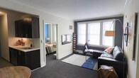 This single-floor space showcases a clean and contemporary design, centered around an open-concept kitchen that serves as the heart of the layout. The kitchen is outfitted with rich, dark cabinetry that provides a striking contrast against the light countertops, creating a bold and modern aesthetic. Stainless steel appliances — including a full-size refrigerator, built-in microwave, and electric range — offer both style and functionality. A generous kitchen peninsula extends into the living area, featuring a double undermount sink with a modern chrome faucet, making it ideal for meal prep and casual dining. The light-toned countertops and neutral wall colors contribute to a bright and airy ambiance throughout the space. Flush-mount ceiling lighting provides consistent illumination, enhancing the clean lines of the interior. The entryway, visible from the kitchen, features a solid door with a door closer, lending a sense of security and practicality. Crown molding details along the ceiling add a subtle architectural touch, elevating the overall finish of the space. The open layout promotes a seamless flow between the kitchen and living areas, making the space feel connected and welcoming.