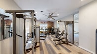 Located at 8965 E Florida Ave in Denver, Colorado, this single-story fitness center offers a comprehensive and thoughtfully designed workout facility. The main gym area features striped hardwood-style flooring in a blend of light and dark tones, recessed lighting, and a large decorative wall mural depicting a dramatic mountain landscape, adding an inspiring, Colorado-appropriate ambiance to the space. A large ceiling fan overhead aids with ventilation and comfort throughout workouts. The main floor is well-equipped with a variety of cardio machines, including treadmills, ellipticals, and a stationary bike, alongside multiple strength-training cable machines, a multi-functional rack, and weight benches. Large windows fitted with sheer roller shades allow natural light to fill the space while maintaining a comfortable environment. An adjacent room provides additional strength training equipment, including a rowing machine, free weights, and a squat rack, finished with a patterned carpet that adds visual distinction. A dedicated studio room features a wall-mounted ballet barre, mirror panels, and a large window overlooking lush greenery outdoors — ideal for stretching, yoga, or barre classes. A connecting hallway leads to an exterior door and provides access to a spa area featuring a corner soaking tub, completing the full wellness offering of this facility.