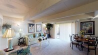 This two-bedroom, one-and-a-half-bath townhouse is spread across two floors, offering a well-organized layout with distinct living and sleeping areas. The main level centers around a spacious living and dining area characterized by exposed ceiling beams and white-painted brick accent walls that lend a rustic yet refined architectural character. Soft carpeting runs throughout the living space, contributing to a warm, comfortable ambiance. A large sliding glass door floods the room with natural light and provides direct access to an outdoor patio area, making the living space feel open and connected to the exterior.

Adjacent to the living area, the kitchen is efficiently designed with light wood cabinetry, a double stainless steel sink, electric range with built-in microwave, and a pass-through window that maintains visual connectivity with the living room. Hardwood-style flooring grounds the kitchen in a practical, easy-to-maintain finish.

The two bedrooms, located on the upper level, each feature the same exposed beam detailing and white brick accent walls seen throughout the home. Both rooms are carpeted and offer ample space for furnishings. The full bathroom includes a tub-shower combination with tile flooring, while a half-bath provides added convenience for guests on the main level.