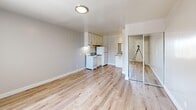 Located at 629 S Main St in downtown Las Vegas, NV, this efficiently designed studio unit offers a thoughtfully organized single-floor layout. The open living area is bright and functional, featuring light hardwood-style flooring that runs seamlessly throughout the entire space, creating a clean, cohesive aesthetic.

The kitchenette is positioned along one wall and is equipped with white cabinetry, an electric range, a full-size refrigerator, and a sink with granite-style countertops. Upper cabinets provide practical storage, and a ventilation hood is installed above the range. Adjacent to the kitchen, a secondary countertop with a sink offers additional workspace or bathroom prep functionality, visible through a partial partition wall.

A dedicated sleeping or closet alcove features full mirrored sliding closet doors that reflect light and contribute to a more open feel within the compact footprint. The main living area also includes a wall-mounted air conditioning unit and a window that allows natural light to enter while offering views of the surrounding neighborhood.

The unit is finished with neutral gray walls, white trim, and flush-mount lighting. The overall design is clean and minimal, making efficient use of the available space. This unit is well-suited for a single occupant seeking a practical, move-in-ready space in a central Las Vegas location.