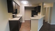 This single-floor residence is designed with functionality and style in mind. The kitchen serves as a central highlight, showcasing rich dark wood cabinetry that contrasts attractively with light-toned countertops. The space is equipped with a full suite of black appliances, including a refrigerator, electric range, built-in microwave, and dishwasher, providing everything needed for comfortable day-to-day living. A peninsula counter extends into the space, creating a convenient breakfast bar or additional prep area while visually separating the kitchen from the adjoining room. The layout follows a galley-style configuration, maximizing efficiency with ample cabinet storage on both sides. Durable hardwood-style flooring runs throughout, adding warmth and easy maintenance underfoot. The ceiling features a textured finish, and recessed ventilation keeps the space well-aired. Neutral wall tones complement the overall color palette, creating a cohesive and inviting atmosphere. A passthrough opening connects the kitchen to an adjacent area, enhancing the sense of flow between spaces. Crisp white baseboards and trim provide a clean finishing touch throughout. The space is well-lit by a ceiling-mounted flush light fixture, ensuring brightness across the full kitchen area.