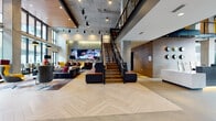 This contemporary lobby and amenity space showcases an industrial-modern design characterized by soaring double-height ceilings with exposed concrete surfaces and warm wood accents. The expansive main lobby is anchored by a striking herringbone-patterned hardwood floor that draws the eye toward a central open-tread staircase with black metal railings, leading to an upper mezzanine level. Track lighting and pendant fixtures illuminate the space, complementing natural light that pours in through floor-to-ceiling windows offering views of the surrounding urban streetscape.

The lounge area is furnished with an eclectic mix of leather sofas, bold accent chairs in jewel tones, and cowhide rugs, creating an inviting, social atmosphere. A large-format TV display serves as a media focal point within this seating zone. Adjacent to the main lobby, a secondary amenity area features sleek white cabinetry with quartz countertops, additional TV displays, and ample natural light. A recessed nook with a tufted bench and side tables offers a more intimate seating option framed by exposed brick. Connecting corridors feature richly toned carpet, concrete ceilings, and curated wall art, tying the overall aesthetic together cohesively.