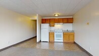 This single-floor unit offers a straightforward and functional layout with distinct living, sleeping, kitchen, and bathroom areas. The open-concept main area combines the kitchen and living space into one cohesive room, with a clear transition between tile and carpet flooring marking each zone. The kitchen is equipped with warm oak cabinetry, granite-patterned countertops, a white electric range with overhead ventilation, a double-basin sink, and a full-size refrigerator. A drop-style ceiling with tile paneling and flush-mount lighting gives the kitchen and living area a clean, uniform appearance. The living space is generously sized, offering ample room for furniture arrangement, with neutral walls and dark baseboard trim throughout. The bedroom is a spacious, carpeted room featuring two windows with red-trimmed frames that let in natural light and provide outdoor views. A wall-mounted HVAC unit and baseboard heating ensure year-round climate comfort. The bathroom retains a classic mid-century character with full pink ceramic wall tile surrounding a soaking tub and shower combination, complemented by mosaic tile flooring and a standard toilet. Overall, the unit presents a practical and livable space with a blend of functional fixtures and retro design elements.