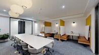Located at 500 W 41st Ave in Denver, CO, this thoughtfully designed collaborative workspace offers a versatile and professional environment suited for both group meetings and individual focus work. The single-floor layout is efficiently organized to support a variety of work styles within a compact yet functional footprint.

At the center of the room sits a long, rectangular white conference table surrounded by ergonomic rolling chairs, making it well-suited for team meetings or presentations. A large flat-screen display is mounted on a textured white pegboard accent wall at one end of the table, accompanied by a trio of elegant globe pendant lights that provide warm, focused illumination overhead.

Along the opposite wall, a row of semi-private booth workstations offers a quieter alternative for focused tasks or smaller conversations. These booths feature warm cognac leather bench seating, wood-framed privacy panels with mustard yellow accents, and individual wall-mounted green pendant lights, creating a cozy yet modern aesthetic.

The space is finished with blue-gray patterned carpet tiles, white walls, and recessed ceiling lighting that complement the overall clean and contemporary design. Glass partition walls near the entrance contribute to an open, airy feel while maintaining visual separation. The cohesive mix of collaborative and semi-private seating makes this space highly adaptable for a range of professional uses.