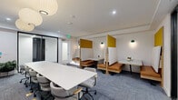 Located at 500 W 41st Ave in Denver, CO, this thoughtfully designed collaborative workspace offers a versatile and professional environment suited for both group meetings and individual focus work. The single-floor layout is efficiently organized to support a variety of work styles within a compact yet functional footprint.

At the center of the room sits a long, rectangular white conference table surrounded by ergonomic rolling chairs, making it well-suited for team meetings or presentations. A large flat-screen display is mounted on a textured white pegboard accent wall at one end of the table, accompanied by a trio of elegant globe pendant lights that provide warm, focused illumination overhead.

Along the opposite wall, a row of semi-private booth workstations offers a quieter alternative for focused tasks or smaller conversations. These booths feature warm cognac leather bench seating, wood-framed privacy panels with mustard yellow accents, and individual wall-mounted green pendant lights, creating a cozy yet modern aesthetic.

The space is finished with blue-gray patterned carpet tiles, white walls, and recessed ceiling lighting that complement the overall clean and contemporary design. Glass partition walls near the entrance contribute to an open, airy feel while maintaining visual separation. The cohesive mix of collaborative and semi-private seating makes this space highly adaptable for a range of professional uses.