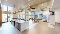 Located at 880 W Euless Blvd in Euless, Texas, this contemporary clubhouse offers a generously sized, single-floor open-concept layout designed for community gathering and recreation. The space immediately impresses with its soaring double-height ceilings, large-format tile flooring, and a sophisticated palette of warm neutrals and natural textures throughout.

At the heart of the clubhouse is a dramatic central living area anchored by a striking textured stone column that rises to the ceiling, serving as both a structural and decorative focal point. Overhead, a curated collection of statement pendant lights — including woven rattan fixtures and illuminated ring chandeliers — add layers of warm, ambient lighting alongside recessed ceiling lights.

A sleek white-quartz island defines the kitchen and entertaining zone, complemented by a full wet bar with bar seating and a mounted television — ideal for social gatherings. A pool table positioned centrally reinforces the recreational character of the space.

Multiple seating arrangements, including plush lounge chairs and accent sofas, are thoughtfully distributed to encourage conversation and relaxation. Floor-to-ceiling black-framed windows along one wall flood the interior with natural light while offering unobstructed views of the outdoor resort-style pool. Curated artwork and greenery add refined, welcoming touches throughout.
