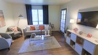 This single-floor space offers a welcoming and well-appointed interior centered around a comfortable living area. The room showcases rich hardwood-style flooring that runs throughout, lending warmth and continuity to the space. Large windows dressed with floor-length navy curtains flood the room with natural light, while a secondary window fitted with wood-slat blinds adds versatility in light control. The living area is thoughtfully arranged with a contemporary grey sofa accented by orange and white throw pillows, creating an inviting pop of color. A sleek glass-and-chrome coffee table sits atop a plush area rug, anchoring the seating arrangement with modern elegance. A clean white shelving unit along one wall provides practical storage and display space, complemented by tasteful décor including greenery and accent pieces. The neutral wall tones create a calm backdrop that allows the furnishings and natural light to take center stage. The overall ambiance is modern, bright, and functional, offering a comfortable and visually cohesive living environment that balances style with everyday practicality.