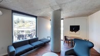 This single-floor space exudes a refined, contemporary aesthetic defined by a striking combination of raw and refined materials. The exposed concrete ceiling lends an industrial edge, while warm hardwood flooring underfoot provides a natural counterbalance, creating a thoughtful tension between the two surfaces. A prominent cylindrical structural column serves as an architectural focal point, anchoring the open-plan layout and adding to the space's bold character.

Floor-to-ceiling windows draw in generous amounts of natural light and frame panoramic views of the surrounding urban and natural landscape, including lush tree canopies and neighboring structures. The seamless visual connection to the outdoors enhances the sense of openness and airiness throughout.

The living area features a sleek, low-profile modular sofa in deep charcoal, complemented by a small accent table, while a dedicated dining zone accommodates a round table with elegant wooden chairs. A sculptural lounge chair adds a design-forward touch near the window. Wall-mounted artwork contributes a gallery-like quality to the interior.

Overall, the space presents a cohesive, architecturally driven environment that balances minimalism with warmth — ideal for those who appreciate modern design with an artistic sensibility.