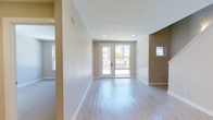 This two-story space is designed with a clean, modern aesthetic that emphasizes openness and natural light. The main living area showcases smooth light hardwood flooring that runs throughout, complementing the neutral, warm-toned walls that create a calm and inviting atmosphere. Recessed ceiling lights provide even illumination, enhancing the airy feel of the room.

A standout feature of the main living area is the set of white-framed French doors that lead directly to an outdoor balcony, allowing abundant natural light to flood the interior. The balcony offers views of the surrounding exterior, adding a sense of connectivity to the outdoors. Windows positioned on adjacent walls further contribute to the well-lit environment throughout the day.

Architectural details such as clean white baseboards, smooth wall transitions, and a subtle accent wall in a deeper taupe tone add visual interest without overwhelming the space. A small pass-through window or niche is visible on one wall, suggesting a thoughtful, functional layout. The two-floor configuration offers additional living space and flexibility, making this a well-appointed and versatile property.