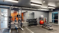 This modern fitness center offers a comprehensive, single-floor workout facility thoughtfully divided into distinct training zones. The space features polished concrete flooring throughout the main areas, transitioning to rubber matting in the strength training sections to provide cushioning and safety during heavy lifts.

The strength training zone is well-appointed with a power rack, a full set of dumbbells on tiered racks, weight plates, and multiple adjustable benches. Full-length wall mirrors line this area, enhancing the sense of space and allowing users to monitor their form. A TRX suspension system is mounted for functional training, and medicine balls and resistance bands add further versatility.

The cardio zone features treadmills, an elliptical, a stationary bike, and a rowing machine, all positioned near large windows that bring in natural light and views of the surrounding brick exterior. A wall-mounted flat-screen TV accompanies this area, set against a bold orange accent wall that adds energetic character to the space.

The facility also includes a separate glass-walled room accessible through white-framed glass doors, ideal for stretching, yoga, or personal training sessions. Industrial-style exposed ductwork runs along the dark textured ceiling, complemented by rows of bright linear LED lighting that illuminate the entire space evenly.