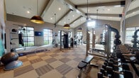This single-level fitness center is designed to accommodate a wide range of workout needs within a thoughtfully organized, open layout. The space is anchored by dramatic vaulted ceilings supported by exposed wood-tone beams, which lend an architectural warmth to the otherwise modern interior. Stylish dome pendant lights hang at varying heights throughout, complementing the recessed lighting and contributing to a well-lit environment.

The flooring features a patterned, multi-tone carpet tile design that subtly delineates different functional zones across the space. Large black-framed windows line multiple walls, flooding the interior with natural light and providing views of surrounding greenery and the exterior landscape.

The strength training area is comprehensively outfitted with a full rack of dumbbells, weight benches, a cable machine, a multi-functional power rack with weight plate storage, and wall-to-wall mirrors that enhance the sense of space. The cardio zone features an array of treadmills, stationary bikes, ellipticals, and a rowing machine, each equipped with individual monitors.

A functional fitness area includes kettlebells, balance tools, stability balls, and a heavy punching bag. One wall is boldly decorated with a large geometric mural in teal, gold, and gray, adding a vibrant focal point. A wall-mounted television and a small refreshment station complete the amenities.