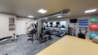 This fitness center is a comprehensively outfitted exercise facility designed to support a wide range of workout routines. The space is laid out on a single floor and features a practical, open floor plan that accommodates multiple zones for different types of training.

The cardio section is prominently featured, housing a row of treadmills equipped with individual monitors, as well as elliptical machines and a stationary bike, allowing users to engage in varied cardiovascular workouts. Adjacent to the cardio area, a multi-station cable and weight machine provides options for full-body strength training, while a dedicated free weights zone includes a tiered dumbbell rack with a wide range of weights, kettlebells, and an adjustable workout bench.

A large wall-mounted mirror spans one end of the room, enhancing the sense of space and allowing users to monitor their form during exercise. The facility also includes a selection of stability and medicine balls stored on a wall-mounted rack, supporting functional fitness and flexibility training.

The flooring combines carpet tiles in the equipment areas with a smooth hardwood-style surface in the open exercise zone. Overhead, exposed ductwork runs alongside recessed panel lighting, giving the space a utilitarian yet clean aesthetic. Windows along the walls allow natural light to supplement the bright overhead lighting, contributing to an energizing workout environment. Wall-mounted televisions are positioned throughout for entertainment during workouts.