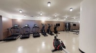 This fitness center is a comprehensively outfitted facility designed to support a full range of workout routines. The space is organized into distinct functional zones, each tailored to a specific type of exercise. The cardio area features a row of treadmills — including Spirit-brand models — alongside elliptical trainers and stair climbers, all equipped with individual digital displays. A row machine provides an additional low-impact cardio option and is prominently positioned in the open floor space. A dedicated cycling area houses multiple stationary bikes with tablet-style screens, set apart by sleek glass-panel partitions with black frames that lend a modern, studio-like feel. The strength training zone includes a cable and pulley multi-functional machine, a flat adjustable bench, and a well-stocked dumbbell rack spanning a range of weights. Floor-to-ceiling mirrors are strategically placed throughout to maximize the sense of space and allow users to monitor their form. The flooring throughout is a light-toned, speckle-finish composite that is both durable and easy to maintain. Flush-mount ceiling lights provide consistent, even illumination across the entire space. The walls are finished in a warm taupe tone, contributing to a clean and focused atmosphere. The glass partitions offer views into an adjacent common area, adding visual connectivity to the broader facility.