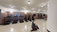 This fitness center is a comprehensively outfitted facility designed to support a full range of workout routines. The space is organized into distinct functional zones, each tailored to a specific type of exercise. The cardio area features a row of treadmills — including Spirit-brand models — alongside elliptical trainers and stair climbers, all equipped with individual digital displays. A row machine provides an additional low-impact cardio option and is prominently positioned in the open floor space. A dedicated cycling area houses multiple stationary bikes with tablet-style screens, set apart by sleek glass-panel partitions with black frames that lend a modern, studio-like feel. The strength training zone includes a cable and pulley multi-functional machine, a flat adjustable bench, and a well-stocked dumbbell rack spanning a range of weights. Floor-to-ceiling mirrors are strategically placed throughout to maximize the sense of space and allow users to monitor their form. The flooring throughout is a light-toned, speckle-finish composite that is both durable and easy to maintain. Flush-mount ceiling lights provide consistent, even illumination across the entire space. The walls are finished in a warm taupe tone, contributing to a clean and focused atmosphere. The glass partitions offer views into an adjacent common area, adding visual connectivity to the broader facility.