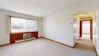 This single-floor unit offers three bedrooms, one bathroom, a kitchen, and a hallway, making efficient use of its compact footprint. The bedrooms feature neutral carpet flooring, white walls, and warm wood trim baseboards that run throughout the unit, lending a consistent and clean aesthetic. The primary bedroom is the most spacious of the three, equipped with a wall-mounted air conditioning unit and large vertical-blind-covered windows that allow natural light and outdoor views. The secondary bedrooms are more modest in size, each with carpeted floors and windows framing exterior views. Closets with hanging rods are found in multiple bedrooms, providing practical storage solutions. The kitchen is outfitted with white cabinetry, a gas range, a refrigerator, a range hood, and a stainless steel sink with countertop workspace — a fully functional setup in a streamlined layout. The bathroom features white tile walls, a wall-mounted sink, a toilet, a bathtub, tile flooring, and a vanity mirror with overhead lighting. Throughout the unit, warm-toned wood door frames and trim provide a cohesive design element, while the textured ceilings and neutral palette create a straightforward, functional living environment.