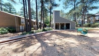 This single-story structure is situated within a lush, wooded setting dominated by tall, mature pine trees that provide generous shade and a tranquil natural atmosphere. The exterior is finished in a neutral gray siding, complemented by a distinctive angled roofline that adds architectural character to the building's profile. A covered patio area is attached to the front of the building, offering a sheltered outdoor space furnished with seating, ideal for relaxing while enjoying the surrounding greenery. A concrete walkway leads up to the entrance, lined with well-maintained landscaping and bordered by a black iron fence that defines the property boundary. The grounds feature a mix of open sandy areas and planted beds, reflecting the natural pine needle ground cover typical of this wooded environment. Additional residential buildings are visible in the background, suggesting this structure is part of a larger planned community. The overall ambiance is calm and secluded, with the dense tree canopy filtering natural light across the exterior and creating a peaceful, park-like setting throughout the property.