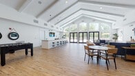 Located at 17151 Windward Blvd in Milford, Delaware, this single-floor community clubhouse is designed to serve as a welcoming and versatile gathering space. The architecture is defined by dramatic vaulted ceilings with exposed white-painted beams that rise to impressive heights, creating an expansive, airy atmosphere throughout the main hall. Warm, light-toned hardwood flooring runs continuously across the space, complementing the crisp white walls and lending a clean, modern-coastal aesthetic.

The main open area is thoughtfully arranged to accommodate multiple uses simultaneously. A central bar counter with industrial-style stools provides a casual social hub, accented by pendant lighting overhead and a mounted flat-screen TV. Nearby, a foosball table adds a recreational focal point, while round dining tables with wooden chairs offer comfortable seating for gatherings and events.

A lounge area features plush blue sofas, side tables, a wall-mounted television, and views directly out to the outdoor pool and patio through large glass doors. Floor-to-ceiling arched windows along the rear wall flood the interior with natural light and frame lush outdoor greenery, seamlessly connecting the interior to the exterior amenity space. Multiple sets of double glass doors provide direct access to the pool area, enhancing the indoor-outdoor flow of this well-appointed community facility.