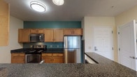 Located at 191 Beach in Salisbury, MA, this single-floor unit offers a thoughtfully designed layout with a comfortable and functional living environment. The kitchen is a standout feature, boasting warm maple cabinetry, dark granite countertops, and a full suite of stainless steel appliances including a refrigerator, range, and microwave. A bold teal accent wall adds a pop of color, while durable hardwood-style flooring grounds the space with warmth. A peninsula counter with pendant lighting connects the kitchen to the open living and dining area, which is finished with plush neutral carpeting and soft, warm-toned walls that create an inviting atmosphere. A sliding glass door allows natural light to fill the main living space and provides access to an outdoor area. The bedrooms are generously sized, carpeted for comfort, and feature large windows that let in ample natural light — one offering views of the surrounding landscape. The bathroom is clean and well-finished, with tile flooring, a vanity with a granite-topped wood cabinet, a large mirror with overhead lighting, and classic white fixtures. Overall, this space combines practical design with welcoming finishes throughout.