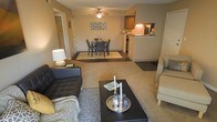 This single-floor residence offers an open-concept layout that seamlessly connects the living, dining, and kitchen areas into one cohesive space. The living area features plush carpeting underfoot and is furnished with a tufted leather sofa complemented by a textured area rug, centered around a dark-finished coffee table with decorative accents. A ceiling fan overhead provides both light and airflow to the space.

The dining area sits just beyond the living room, accommodating a round table with seating for four, positioned atop a lighter area rug that helps define the zone. Neutral-toned walls and warm lighting contribute to a cozy, inviting ambiance throughout.

The kitchen is accessible via a raised breakfast bar counter, creating a natural visual boundary while maintaining the open feel of the floor plan. Warm wood cabinetry and overhead lighting give the kitchen a functional and welcoming character. A hallway visible in the background suggests additional rooms, including what appears to be a closeted storage area. The overall design balances practicality with a comfortable, livable aesthetic.