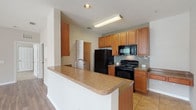 This single-floor residence offers a well-organized layout that balances functionality and comfort throughout its living spaces. The kitchen serves as a central feature, outfitted with warm-toned wood cabinetry, granite-style countertops, and a full suite of black appliances including a refrigerator, range, and built-in microwave. A tiled backsplash adds a decorative touch, while the peninsula breakfast bar provides additional counter space and a natural transition into the main living area. A built-in desk area with matching cabinetry offers a practical workspace within the kitchen zone. Tile flooring runs through the kitchen and extends into a dedicated laundry room, which houses a full-size washer and dryer. The living room is bright and open, featuring durable wood-style flooring, a ceiling fan with light fixture, and large windows that allow generous natural light. Sliding glass doors with vertical blinds open to an outdoor balcony with exterior views. The bedroom is carpeted for comfort, includes a ceiling fan, and is lit by a sizable window. The bathroom is finished with tile flooring, a granite-topped vanity, and a tiled soaking tub and shower combination.