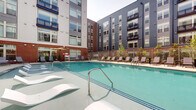 Located at 7970 Maitland St in McLean, Virginia, this outdoor amenity space centers around a generously sized resort-style swimming pool set within a well-designed courtyard. The pool area is enclosed by a multi-story building whose façade blends contemporary gray paneling with classic brick accents, large windows, and private balconies that overlook the space below. The pool itself features a smooth, curved design with striking in-water loungers positioned along a shallow ledge — ideal for relaxing at the water's edge. A wide, open pool deck surrounds the water and is outfitted with rows of modern sling-style lounge chairs and side tables, providing ample space for sunbathing and leisure. Mature greenery and ornamental plantings are thoughtfully integrated along the perimeter, softening the architectural lines and adding a natural element to the setting. A lifeguard stand and shaded umbrella seating area are positioned along one side, complementing the safety and comfort features of the space. The courtyard orientation allows for open sky views while maintaining a sheltered, intimate atmosphere. The overall design reflects a polished, upscale aesthetic that prioritizes outdoor living and community recreation.