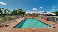 This outdoor pool area is a well-appointed recreational space designed for leisure and relaxation. At its center sits a generously sized rectangular swimming pool with clear turquoise water, framed on all sides by warm-toned brick paver decking that provides ample room for lounging and circulation. The pool is accessible via stainless steel entry rails and features clearly marked depth indicators along its edges.

The surrounding deck is furnished with multiple chaise lounges positioned to take advantage of open sky views, along with colorful Adirondack-style chairs in vivid hues that add a casual, welcoming character to the space. A pair of patio umbrella tables on one side of the deck offer shaded seating for gathering and socializing.

A dedicated pool house structure anchors one end of the area, featuring a pitched shingle roof, a covered entryway, and white-painted exterior walls. This building likely provides restroom facilities and storage support for the pool. The entire area is enclosed by a white metal vertical-picket fence, providing both safety and a clean aesthetic boundary. Mature trees visible beyond the fence create a natural green backdrop, enhancing the open-air ambiance of the space.