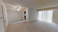 This single-floor residence offers a straightforward and functional layout designed to maximize comfort and usability. The main living area is generously proportioned, featuring wall-to-wall plush carpeting that adds warmth underfoot throughout the space. A ceiling fan with an integrated light fixture is centrally mounted, providing both overhead lighting and air circulation to keep the area comfortable year-round. One wall is dominated by a large sliding glass door dressed with vertical blinds, allowing an abundance of natural light to fill the room while offering access to the outdoors. The neutral, light-toned walls create a clean and versatile backdrop, lending the space a bright and open ambiance. A partial dividing wall defines the transition between the living area and the rest of the unit, adding a subtle architectural element to the open floor plan. Directly accessible from the living space is a compact kitchen, visible through an open doorway and featuring dark cabinetry and essential appliances. The overall design emphasizes a simple, practical aesthetic, making it well-suited for everyday living. The combination of natural light, soft flooring, and an open layout gives the space an inviting and easy-to-personalize character.