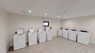 This shared laundry facility offers a clean and functional environment designed to accommodate multiple users simultaneously. The room features several top-loading washing machines and matching dryers arranged in an organized layout, providing ample capacity for residents or guests. The space is finished with light hardwood-style flooring that complements the neutral, off-white walls, creating a bright and welcoming atmosphere. Recessed ceiling lights illuminate the room evenly, ensuring excellent visibility throughout. A small window centered on the back wall adds a subtle architectural detail while allowing a glimpse of natural light. A wall-mounted monitor is positioned near the window, likely serving as a payment or information display for machine use. The machines are uniformly white, contributing to the room's clean, cohesive aesthetic. A security camera is also visible, providing an added layer of safety and peace of mind for users. The single-floor layout ensures easy accessibility, and the open floor plan allows for comfortable movement between machines. Overall, the facility is well-maintained and efficiently designed to meet everyday laundry needs.