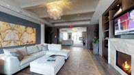 This refined lounge presents a masterclass in contemporary interior design, offering a thoughtfully curated environment ideal for relaxation and socializing. The space is anchored by a stunning bubble-form chandelier that serves as an artistic centerpiece, casting a warm, ambient glow across the room. Underfoot, a rich herringbone-patterned hardwood floor adds depth and visual texture, grounding the sophisticated aesthetic throughout.

The main seating area features a generous white sectional sofa accented with decorative cushions, complemented by a large upholstered ottoman that doubles as a coffee table — currently styled with coffee table books. A bold abstract artwork in warm amber and cream tones adorns one wall, providing an artistic focal point against the deep, neutral-toned walls.

The lounge flows seamlessly into an adjacent gathering area visible through a wide open passage, where large windows invite abundant natural light and floor-length drapery frames the openings with an elegant softness. Lush potted tropical plants are strategically placed throughout, introducing organic warmth and vibrancy to the space. Subtle recessed lighting, coffered ceiling details, and warm wood-toned accents on doors and walls complete the polished, welcoming atmosphere of this well-appointed lounge.