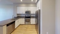 Located at 19401 Parthenia St in the Northridge neighborhood of Los Angeles, this single-floor unit offers a well-designed, contemporary living space with a cohesive neutral color palette and modern finishes throughout. The open-concept layout seamlessly connects the living area to the kitchen, creating a spacious and functional flow. Light wood-look flooring runs through the main living spaces, complemented by recessed lighting and clean white baseboards that lend a crisp, polished appearance. The kitchen is well-appointed with white shaker-style cabinetry, dark countertops, and a full suite of stainless steel appliances, including a refrigerator, electric range, over-the-range microwave, and dishwasher. A peninsula counter further defines the kitchen while maintaining visual openness to the living area. The bedroom features plush carpeting and a window with blinds that allows natural light to filter in while offering privacy. The living room similarly benefits from a large window overlooking greenery, enhancing the sense of connection to the outdoors. Throughout the unit, neutral gray-toned walls provide a versatile backdrop, and the overall design is move-in ready with a clean, modern aesthetic suited for comfortable everyday living.
