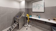 Located at 1989 Cheshire Bridge Rd NE in Atlanta, Georgia, this dedicated pet grooming facility — known as the Wash & Wag — is a thoughtfully designed, single-floor space built to provide a clean, comfortable, and fully equipped environment for pet care. The interior is finished with large-format grey stone-look wall tiles extending halfway up the walls, paired with a mosaic grey tile floor, lending the space a modern and polished aesthetic that is both stylish and easy to maintain. The centerpiece of the grooming area is a professional-grade stainless steel pet wash tub with built-in steps for easy pet access, accompanied by a flexible spray hose and a standalone professional blow dryer. A long white wall-mounted shelf provides ample space for grooming products, sprays, and accessories. A large-panel glass enclosure with sleek black framing houses an additional grooming or drying area, complete with recessed lighting overhead. Elevated pet feeding bowls are conveniently positioned nearby. The space is access-controlled and includes posted Wash & Wag rules for residents. Charming canine-themed wall art adds a welcoming, playful touch. The clean lines, neutral palette, and purpose-built features make this an exceptional amenity for pet owners.