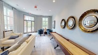 The Second Clubhouse is a thoughtfully designed single-floor amenity space that blends leisure, socializing, and productivity into one cohesive environment. The interiors are finished with light wood-tone flooring laid in a herringbone pattern, soft gray walls, and recessed lighting that creates a bright, welcoming atmosphere throughout.

The main gathering area serves as the social heart of the clubhouse, anchored by a full-length shuffleboard table crafted from rich hardwood. A set of three decorative convex mirrors with ornate gold frames line the adjacent wall, adding a refined, artistic touch. Comfortable cream-toned sofas and plush blue accent chairs are arranged on a patterned area rug, providing ample seating for casual socializing or game play. Large windows and glass doors along one wall invite generous natural light and offer views of the surrounding greenery outdoors.

A secondary lounge area features additional seating alongside a wall-mounted television and a built-in wet bar with marble countertops and a beverage refrigerator — ideal for entertaining. Bold abstract artwork and decorative accents contribute to the space's contemporary aesthetic.

A dedicated workspace is also available, featuring a live-edge wood conference table surrounded by gray upholstered chairs. A vibrant living plant wall installation provides a striking natural focal point, while tall curtained windows keep the area bright and inspiring.