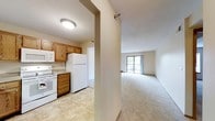 This single-floor space offers a practical and comfortable layout designed for everyday living. The kitchen is equipped with warm oak cabinetry, a white refrigerator, an electric range, and a dishwasher, all set over easy-to-clean tile flooring. A built-in microwave and ample cabinet storage add to the kitchen's functionality. The kitchen flows naturally into the open living area, which is finished with plush wall-to-wall carpeting that adds warmth underfoot. Large sliding glass doors at the far end of the living space flood the room with natural light and provide direct access to a private balcony, offering a pleasant outdoor retreat. The transition between the kitchen and living area is defined by a structural column that subtly separates the two spaces while maintaining an open, airy feel throughout. Neutral wall tones throughout the unit create a clean, versatile backdrop suitable for a variety of décor styles. The overall layout is efficient and well-considered, making the most of the available space while ensuring a comfortable and welcoming ambiance from entry to balcony.
