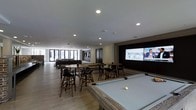 This single-floor entertainment space is thoughtfully designed to serve as a social and recreational hub. The open-plan layout is anchored by a pool table positioned prominently near the center of the room, providing an immediate focal point upon entry. Adjacent to the pool table, several bar-height tables paired with dark wooden stools offer casual seating ideal for gathering and socializing.

One wall features a large projection or display screen framed within a dark recessed panel, creating a dedicated media viewing area well-suited for watching sports or events. The screen is complemented by recessed ceiling lighting throughout the space, ensuring even illumination across all activity zones.

Toward the far end of the room, floor-to-ceiling glass doors and windows flood the space with natural light and provide views to an exterior area, contributing to an open and airy atmosphere. A lounge seating area with sofas is visible in this naturally lit zone, offering a more relaxed retreat within the same space.

The light wood-toned flooring, neutral wall tones, and clean-lined finishes give the space a contemporary, polished aesthetic that balances comfort with modern design sensibility.
