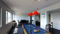 This single-floor recreational space is designed with entertainment and social interaction in mind. The room is anchored by a full-size pool table dressed in blue felt, positioned prominently beneath a striking three-shade pendant light fixture with bold red lamp shades suspended on a chrome bar — a clear focal point that adds a vibrant pop of color to the otherwise neutral interior.

The textured white ceiling and light grey walls create a clean, understated backdrop that keeps the space feeling open and bright. Natural light filters in through windows along one side of the room, contributing to an airy atmosphere.

Adjacent to the pool area, a dining or bar-height table is surrounded by dark chairs, offering a comfortable space for casual seating and socializing. A smaller hanging light with a yellow shade marks this secondary zone, creating a warm, distinct ambiance separate from the main game area.

Framed artwork on the walls adds a touch of character, while a hallway visible in the background suggests connectivity to additional areas. The carpeted flooring ties the functional zones together cohesively, making this a well-rounded space suited for leisure and group gatherings.