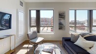 Located at One Chestnut Place in Quincy, MA, this single-floor residence features a thoughtfully designed open layout with contemporary finishes throughout. The living area is anchored by expansive floor-to-ceiling windows that frame sweeping views of the surrounding Quincy streetscape, flooding the space with natural light. Hardwood-style flooring runs through the main living area, complementing the clean, neutral color palette.

The kitchen is outfitted with white cabinetry extending to the ceiling, a gray tile backsplash, stainless steel appliances including a double-oven range, built-in microwave, and French door refrigerator, along with ample counter space and a breakfast bar for casual dining.

Multiple bedrooms feature plush carpeting, soft neutral walls, and large windows offering urban views. The primary bedroom benefits from expansive glazing that maximizes natural light. A carpeted hallway connects the private areas of the residence.

Two bathrooms serve the space: one features a glass-enclosed walk-in shower with gold hardware and large-format tile flooring, while the second offers a soaking tub with subway tile surround and a white vanity. Together, these elements create a cohesive, modern living environment with a bright and airy ambiance.