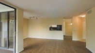 This single-floor space offers an open-concept layout highlighted by rich, dark hardwood flooring that extends throughout the main living area. The living room is spacious and unobstructed, featuring a textured ceiling and a set of adjustable track lights mounted on the ceiling, providing directed illumination across the room. Warm beige walls contribute to a neutral and inviting ambiance throughout.

A defining architectural feature is the pass-through kitchen window, which connects the kitchen to the living area. This opening reveals a glimpse of dark cabinetry and a sink, facilitating a sense of connectivity between the two spaces. The kitchen itself appears to be a separate but accessible area, with a pendant light visible through the adjoining hallway or dining space, adding a subtle decorative touch.

White baseboards provide a clean contrast against the warm-toned walls and flooring. The overall layout is practical and straightforward, with clear sightlines from the main living area into adjoining spaces. The design balances functionality with a comfortable, neutral aesthetic, making the space adaptable for a variety of uses.