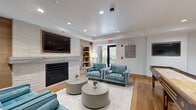 Located at 45 East Newton Street in Boston's South End neighborhood, this thoughtfully designed community lounge offers a well-appointed single-floor layout that balances comfort, entertainment, and functionality. The living and lounge area serves as the heart of the space, anchored by a striking white stacked-stone fireplace wall with a wall-mounted flat-screen TV above the mantle and integrated built-in shelving. Warm hardwood flooring runs throughout, complementing the neutral tones and natural wood accents that define the interior aesthetic. Recessed lighting illuminates the open layout, while a glass door allows natural light to filter in and provides access to the outdoors. Comfortable teal upholstered seating surrounds round coffee tables, creating an inviting atmosphere for gathering. A full-length shuffleboard table and high-top bar seating with white countertops add a recreational dimension to the space. The kitchenette features wood cabinetry, white quartz countertops, a stainless steel sink, microwave, and refrigerator, set against a textured tile backsplash. Glass-paneled sliding doors connect to an adjacent area with a conference or dining setup, extending the versatility of the space for both casual and organized gatherings.