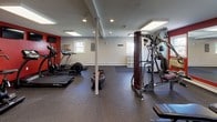 This fitness center offers a thoughtfully organized workout space designed to support a wide range of exercise routines. The interior features durable rubber flooring throughout the main workout area, providing both safety and comfort during exercise. Bold red accent walls add energy to the space, complemented by neutral gray walls and a low ceiling with recessed lighting and flush panel fixtures that keep the area well-illuminated.

The cardio section is equipped with multiple treadmills, an elliptical trainer, a stationary air bike, and rowing machines, with wall-mounted televisions positioned for convenient viewing during workouts. A large mirror along one wall enhances the sense of space and allows for form-checking during exercise. The strength training area includes a multi-station cable machine, a flat weight bench, and a dedicated free weights section featuring a rack of kettlebells and dumbbells in various sizes.

A water dispenser is conveniently located near the strength training area. The entry zone transitions from carpeted flooring to the rubber workout surface and includes a small cubby storage unit, a coat hook rail, and a fire extinguisher. Windows along the perimeter bring in natural light, and a security camera ensures the safety of the facility. The overall layout is open, functional, and designed to accommodate multiple users simultaneously.