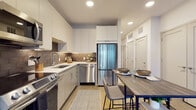 This single-floor unit showcases a thoughtfully designed open layout that connects the kitchen, dining, and living areas into one cohesive space. The kitchen is outfitted with floor-to-ceiling white upper cabinetry, contrasting grey lower cabinets, and a textured tile backsplash that adds depth and warmth. A full suite of stainless steel appliances—including a French door refrigerator, dishwasher, range, and microwave—ensures a fully functional cooking environment. White quartz countertops provide ample workspace, complemented by an undermount sink with a modern pull-down faucet. Light wood-tone flooring runs throughout, unifying the entire space. The dining area accommodates a rustic wood high-top table with upholstered bar-height chairs, positioned conveniently between the kitchen and living room. The living area benefits from a large window that invites natural light and offers an outdoor view, creating a bright and welcoming atmosphere. Recessed lighting throughout provides consistent illumination. The bathroom features wood-look flooring, an earthy olive-green accent wall, and clean white fixtures, offering a calm and stylish retreat. Closet storage is accessible nearby, adding to the unit's functional appeal.