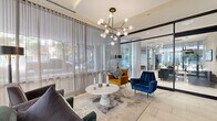 Located in Chicago's vibrant West Loop neighborhood, this ground-floor amenity space at 922 W Washington Blvd offers a generously sized, single-level layout designed for both functionality and refined comfort. The entry lobby immediately impresses with floor-to-ceiling sheer drapery that filters abundant natural light from street-facing windows, large-format white tile flooring, and a striking sputnik chandelier suspended above a curated seating arrangement of velvet accent chairs in rich navy, teal, and mustard tones centered around a marble-top table. A glass-partitioned leasing office sits adjacent to the lobby, offering transparency while maintaining a professional atmosphere. The hallway extends the full length of the building, featuring patterned carpet, recessed and pendant lighting, and exposed cylindrical concrete columns that add an industrial-chic character. A dedicated game and social lounge area includes a pool table, foosball table, built-in media wall with flat-screen television, and a stylish bar-height counter with navy upholstered stools beneath pendant lights. Glass railings and floor-to-ceiling windows provide views to an adjacent outdoor area. Frosted glass-enclosed rooms offer additional private spaces. Throughout, the design balances contemporary elegance with approachable, community-focused amenities.