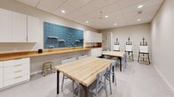 Located in the heart of downtown Minneapolis at 113 S Sixth St, this dedicated art studio is a thoughtfully designed, single-room creative workspace. The room is finished with neutral warm-gray walls, a drop tile ceiling with recessed lighting, and light-toned flooring, creating a clean and well-lit environment conducive to focused creative work.

The studio is furnished with two substantial butcher block worktables paired with gray chairs, offering ample surface area for group projects or individual studio sessions. Multiple black artist easels with canvas boards are positioned throughout the space, indicating its suitability for painting and drawing activities.

Along one wall, a bold teal pegboard display panel with adjustable wooden shelving provides organized storage and display for art tools and supplies. Adjacent to it, a continuous butcher block countertop spans the length of the wall, complemented by white cabinetry above and below, a utility sink, and open shelving units ideal for storing art materials. A white cubby storage unit offers additional organized space for supplies and portfolios.

A dark area rug near the sink zone defines the utility area, while open wall space provides flexibility for displaying or projecting work. The studio is a highly functional, well-appointed space suited for classes, workshops, or individual artistic pursuits.
